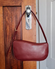 A dark red cactus leather shoulder bag with a curved top edge, crescent-shaped body and a single shoulder strap with a buckle